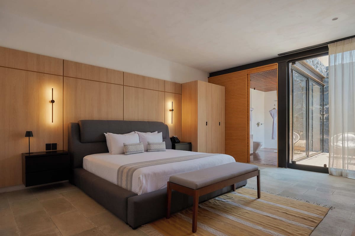 A modern bedroom with a large bed, bedside tables, a bench, and a sliding glass door leading to a balcony.