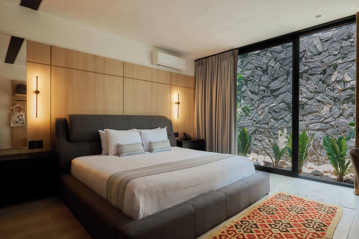 A modern bedroom with a large bed, bedside lamps, a stone wall, and a view of greenery.