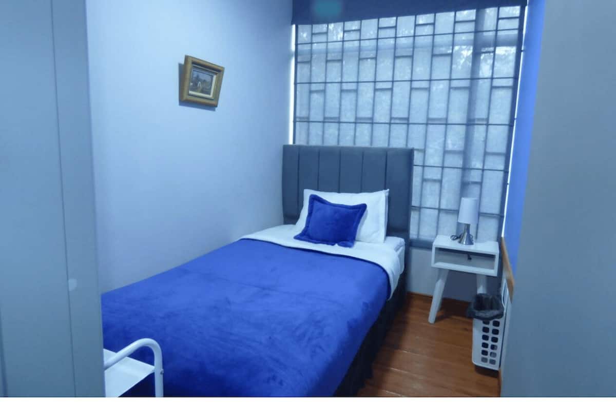 A small, neatly arranged bedroom with a single bed, a bedside table, a nightstand, and a window with a view.