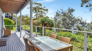 En overdækket terrasse med et bord med glasplade og fletstole, omgivet af frodigt grønt og træer.