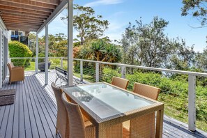 A covered outdoor deck with a glass-top table and wicker chairs, surrounded by lush greenery and trees. - Salt and Serenity (Nelson Bay)