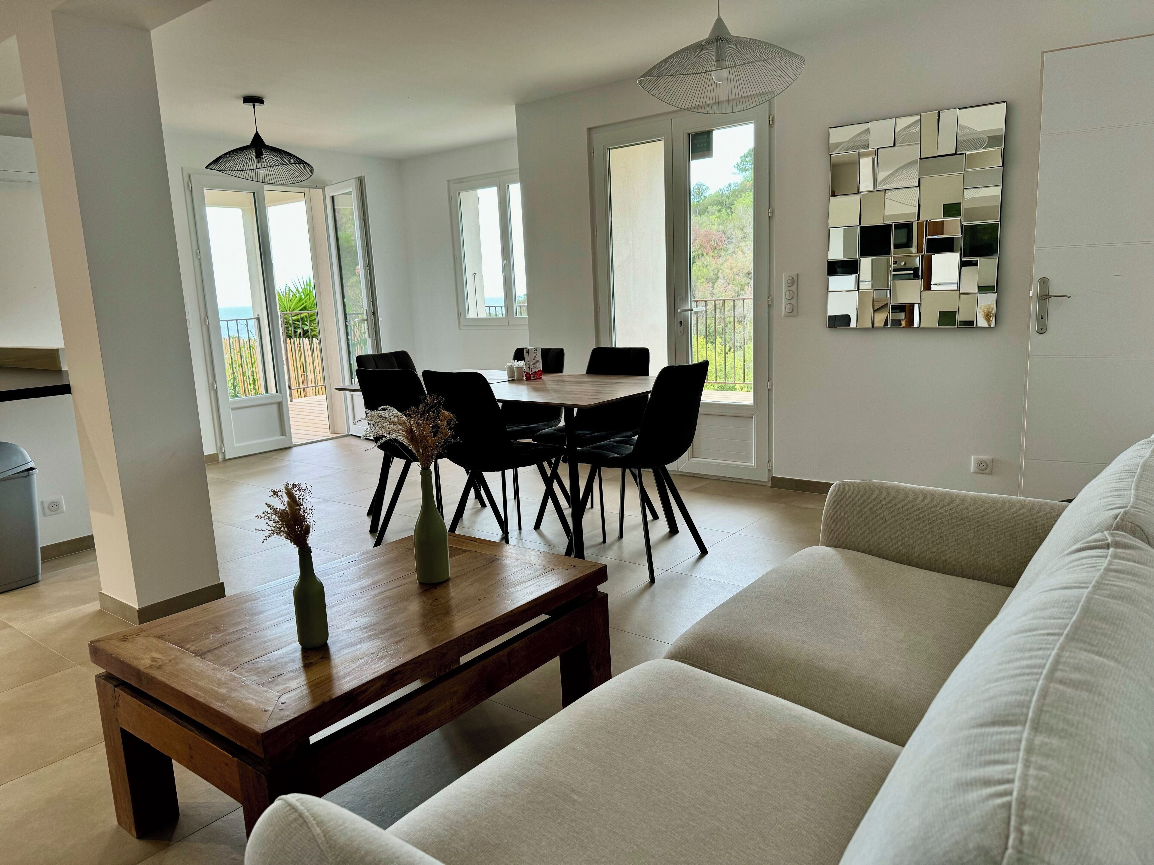 A modern living room with a sofa, a wooden coffee table, and a dining area with a table and chairs. There is a large window with a view of greenery outside.