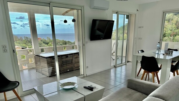 Un salon moderne avec un canapé, une table basse et un coin repas avec des chaises. Un téléviseur est fixé au mur et de grandes portes vitrées offrent une vue sur la mer.