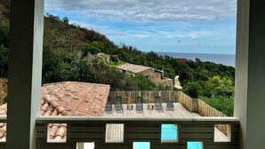 Une terrasse offrant une vue sur un paysage côtier, comprenant une plateforme en bois et un bâtiment en pierre surmonté d’un toit en tuiles.