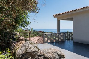 A rooftop terrace with a view of the sea, a white building, and a stone wall.