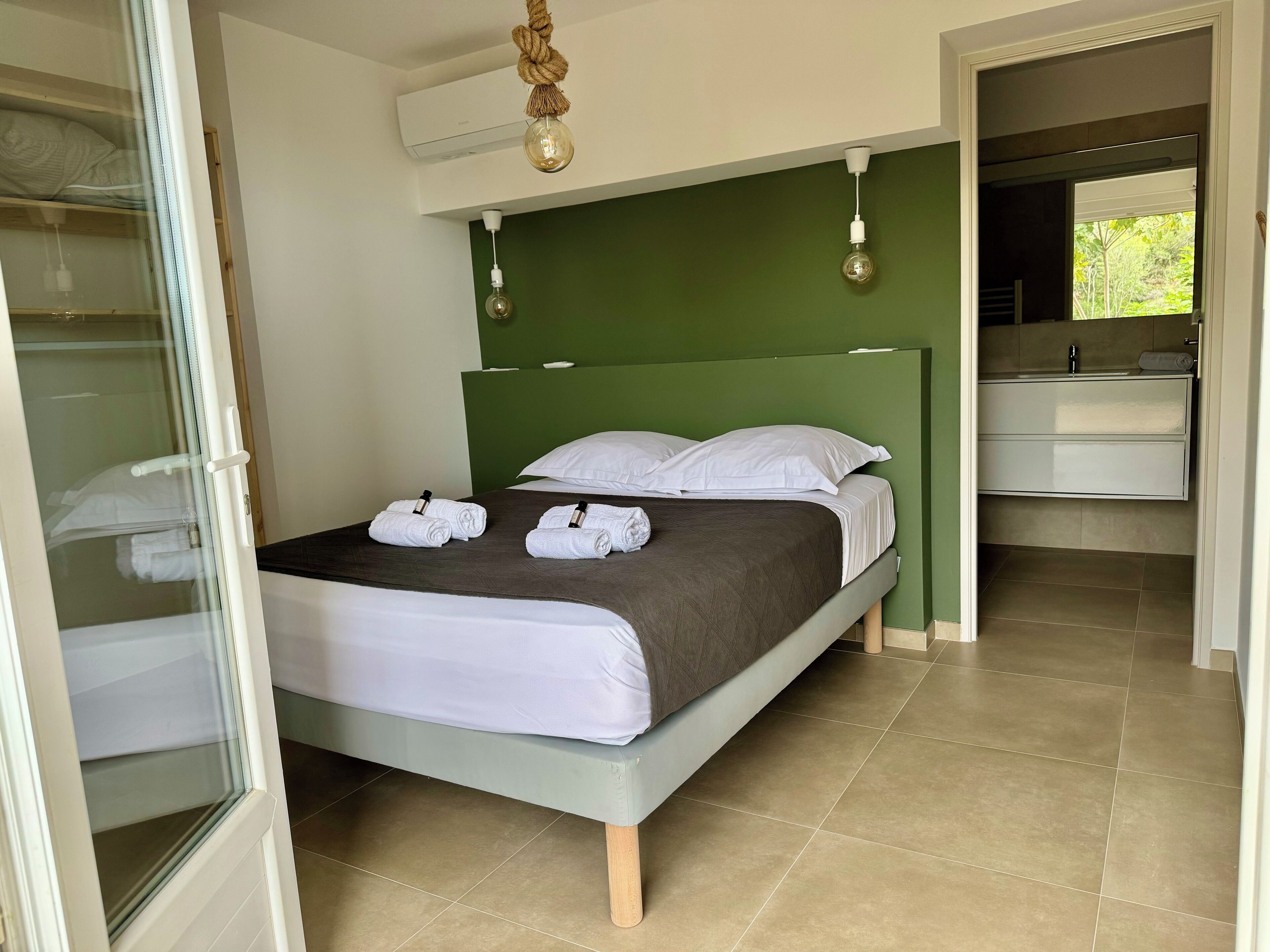 Cama bem arrumada com lençóis brancos e duas toalhas, cabeceira verde e luminária moderna.