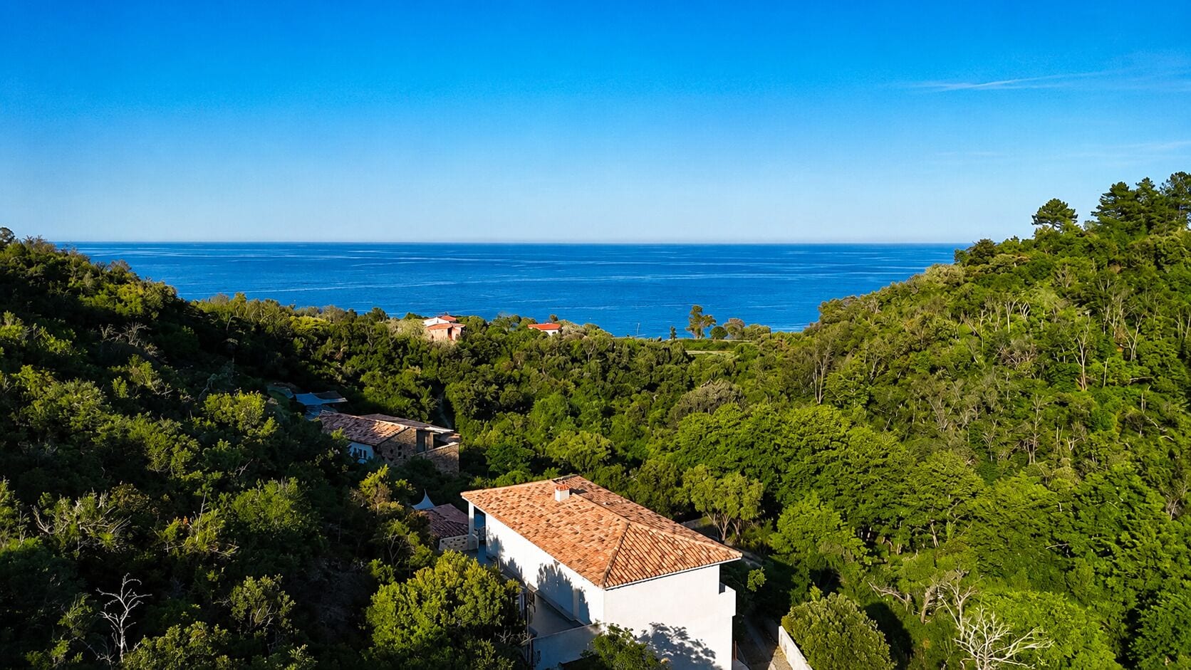 A coastal village with white buildings and red roofs, surrounded by dense greenery and the sea in the background.