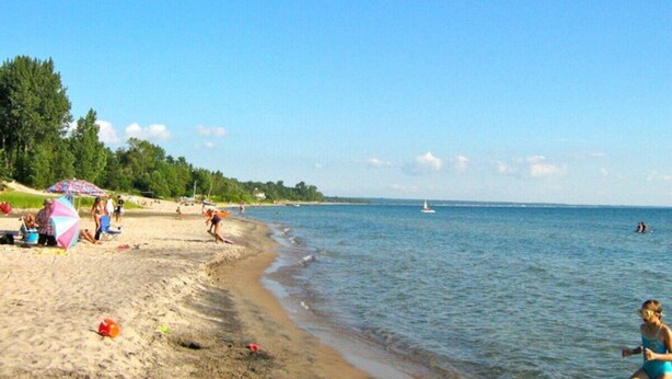 Una spiaggia con persone che prendono il sole e nuotano, una barca a vela all'orizzonte e un cielo azzurro limpido.