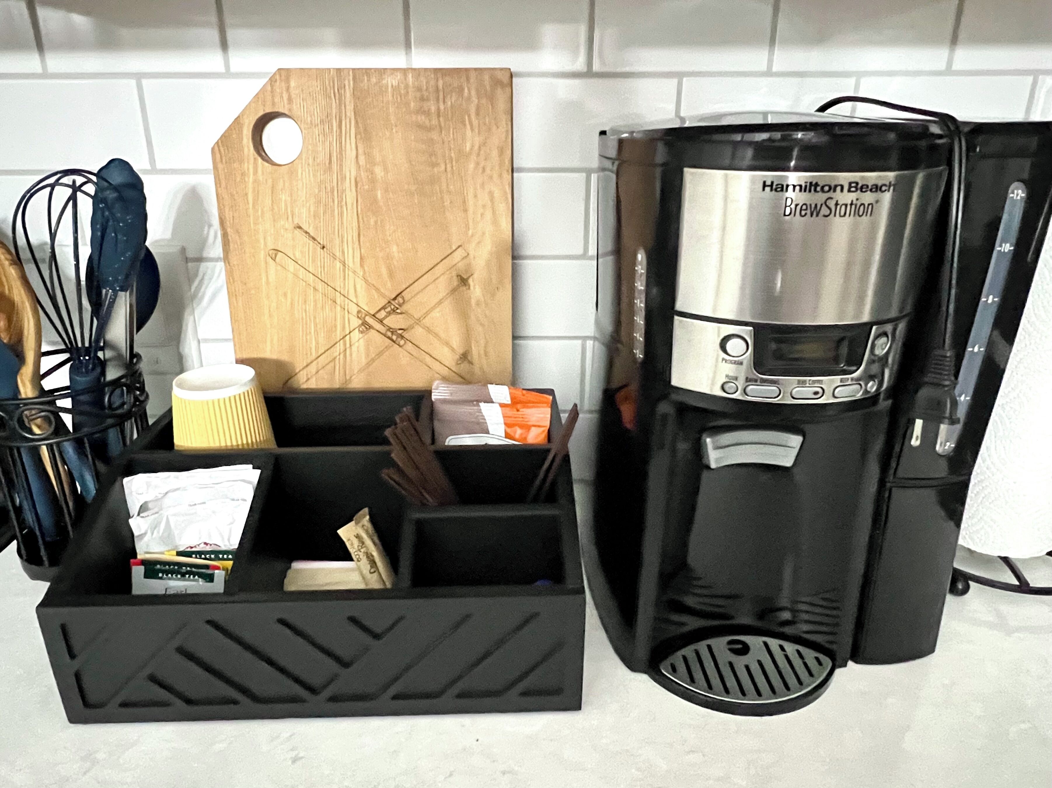 A kitchen with a Hamilton Beach BrewStation coffee maker, a wooden cutting board, and a black storage box containing kitchen utensils and tea boxes.