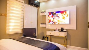 A hotel room with a bed, a TV mounted on the wall, a desk with a coffee maker, and a window with blinds. - 989 Apartments (Abuja)