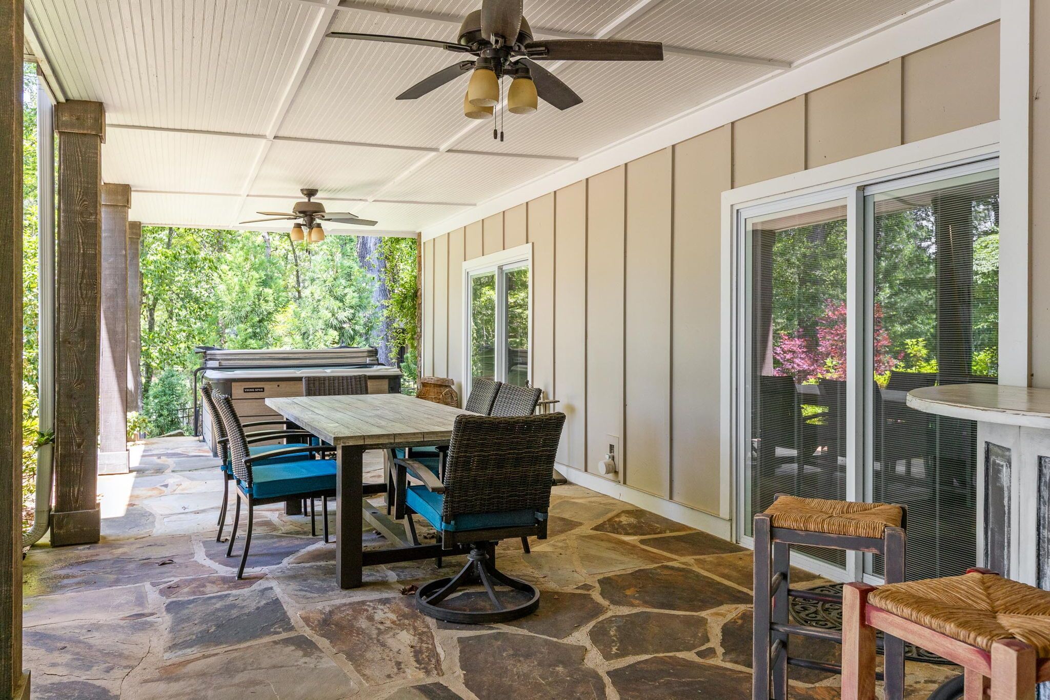 A covered patio with a stone floor, a table and chairs, a ceiling fan, and a view of greenery outside.