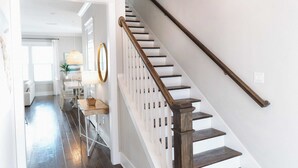 Una escalera con barandas de madera y balaustres blancos que conduce a un espacio interior bien iluminado, con un comedor y una cocina a la vista al fondo.