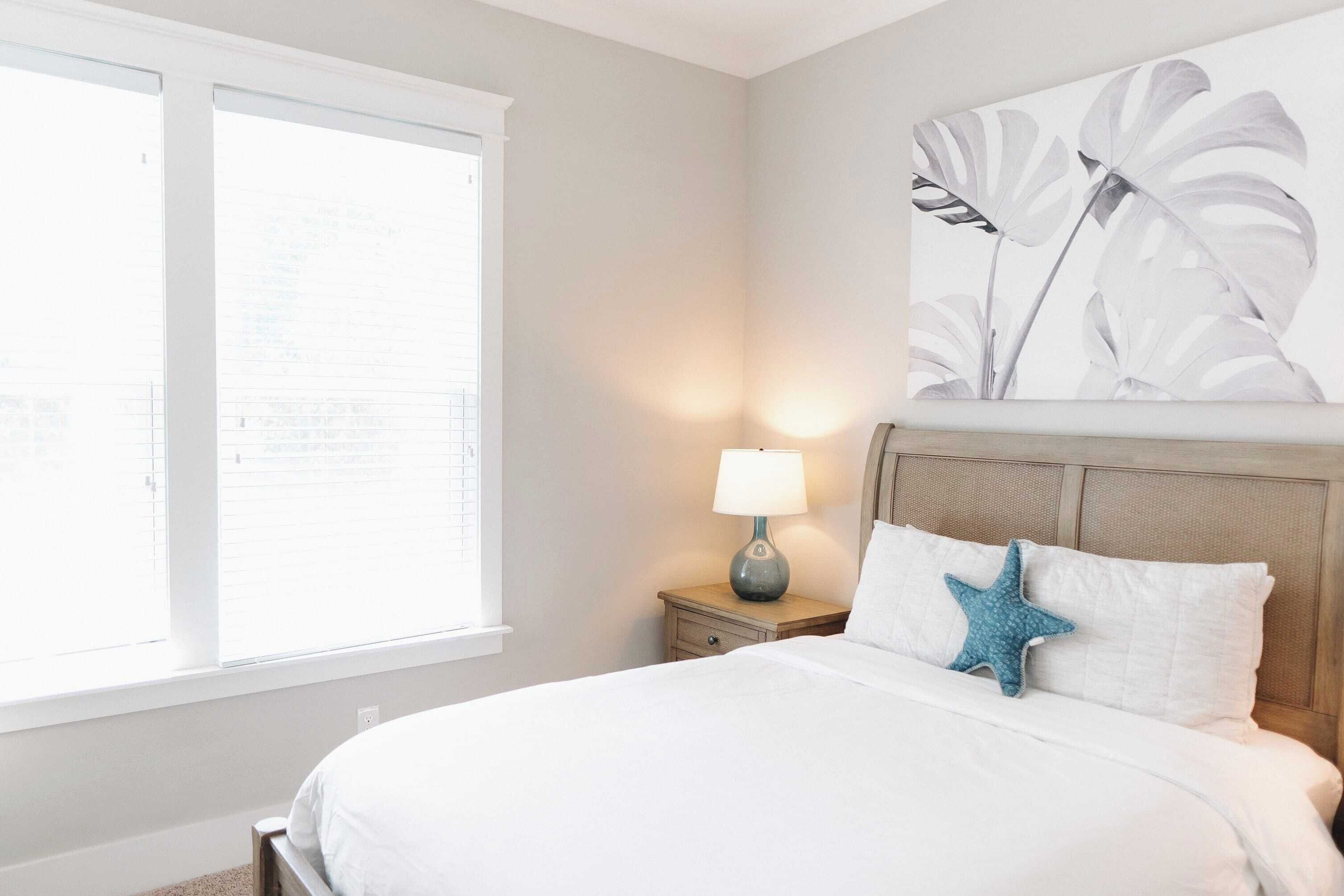 A bedroom with a bed, a nightstand with a lamp, a window, and a wall with a plant painting.