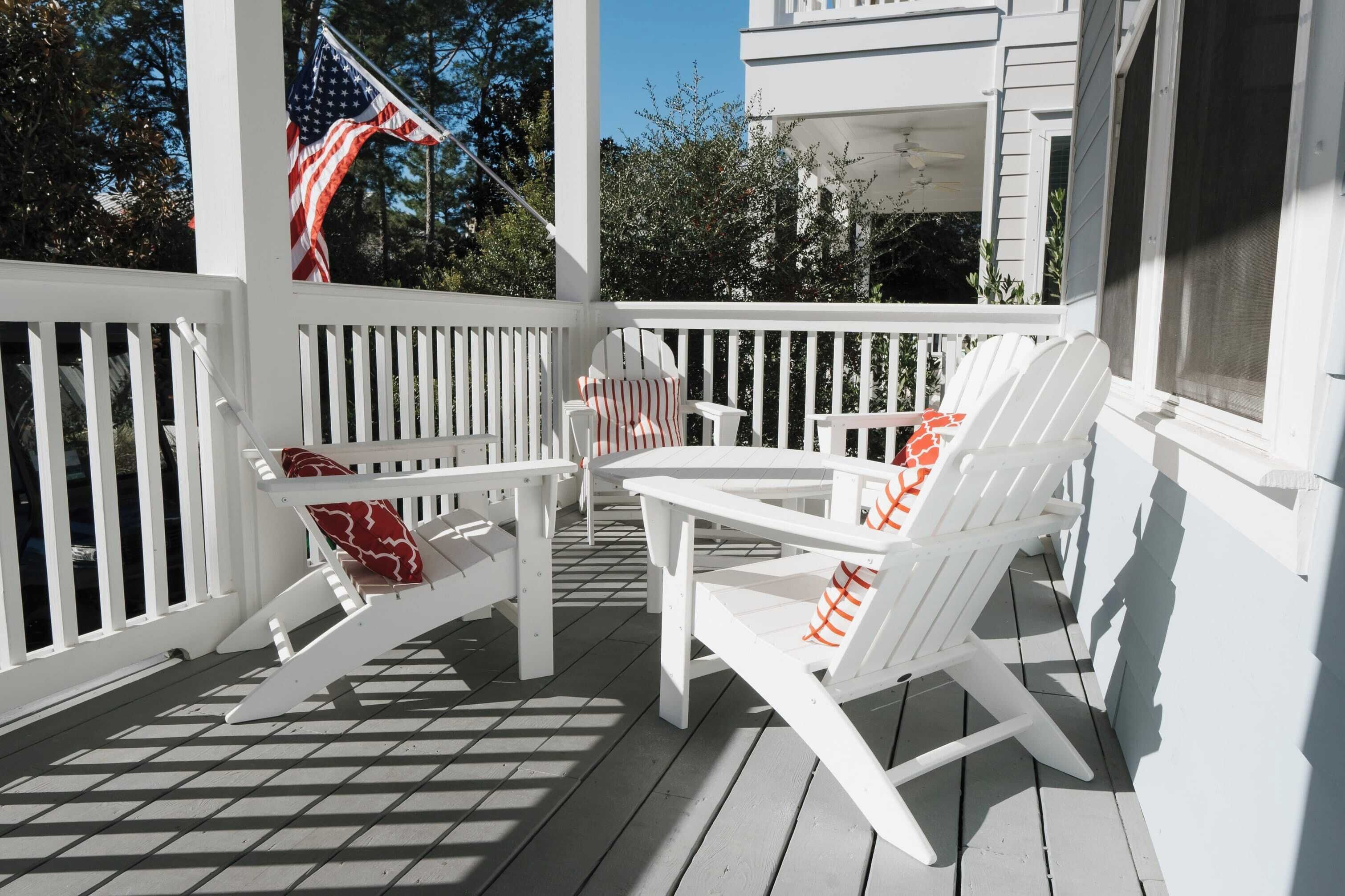 A white wooden deck with two Adirondack chairs and a small table, a flag, and a building in the background.