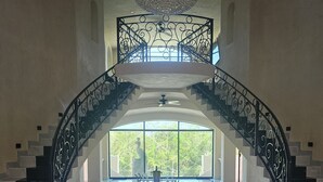 Una gran escalera con barandillas negras y un candelabro, que conduce a un balcón con vistas a la vegetación.