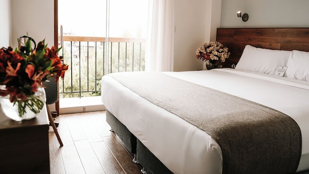 A hotel room with a large bed, a wooden headboard, a window with curtains, and a vase with flowers on a nightstand. - MURU HOMELY HOTEL (Cusco)