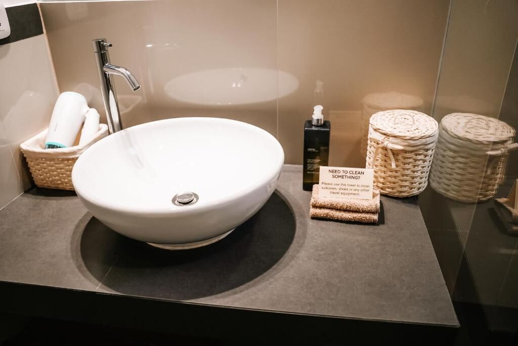 Un baño moderno con un lavamanos blanco grande, un grifo plateado y un espejo. Hay cestas con artículos de aseo y un letrero que dice '¿NECESITA LIMPIAR ALGO?'.