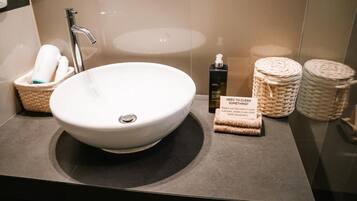 Un baño moderno con un lavamanos blanco grande, un grifo plateado y un espejo. Hay cestas con artículos de aseo y un letrero que dice '¿NECESITA LIMPIAR ALGO?'.