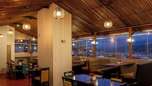 A restaurant with a view of the city at dusk, featuring wooden ceiling beams, hanging pendant lights, and a mix of booth and table seating. - MURU HOMELY HOTEL (Cusco)