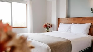 A neatly made bed with white linens and a wooden headboard, a vase of flowers on a bedside table, and a window with curtains. - MURU HOMELY HOTEL (Cusco)
