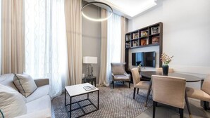 A modern hotel room with a sofa, armchairs, a coffee table, a TV, and a bookshelf.