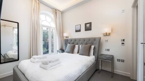 A neatly made bed with white linens and towels, a grey upholstered headboard, and a bedside table with a lamp. - PBC | Two Bedroom Apartment in Kensington - KC:6 (London)