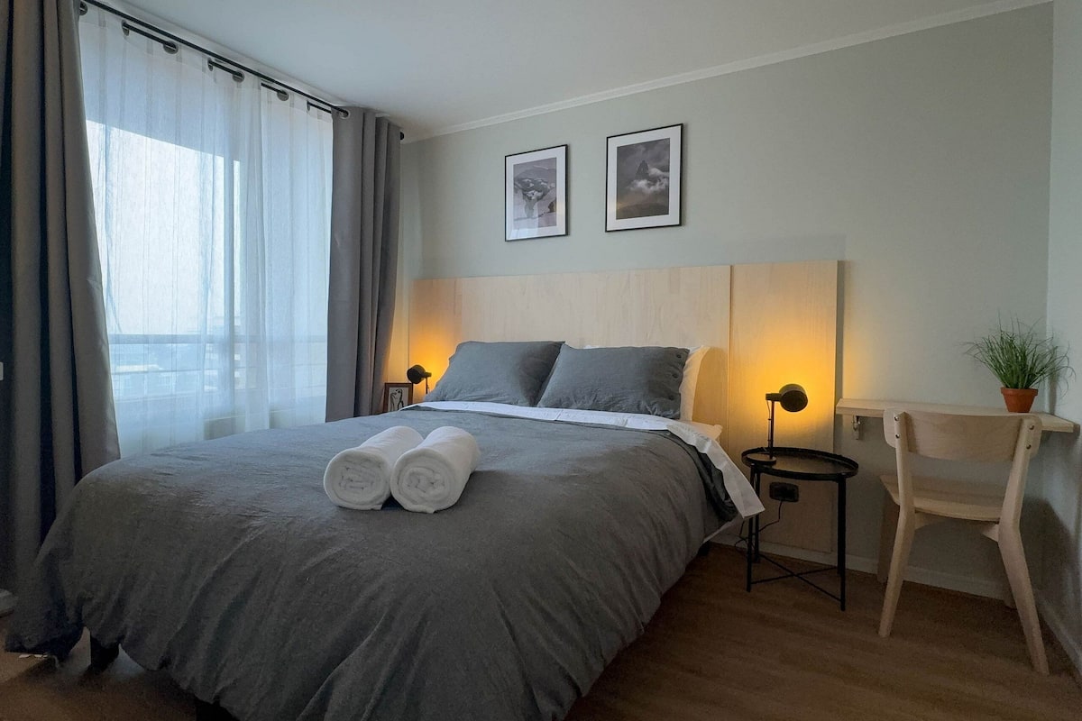 Una cama bien hecha con ropa de cama gris y dos toallas enrolladas, una mesita de noche con una lámpara, una silla y dos cuadros en la pared.