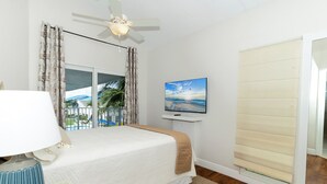 Un dormitorio con cama, televisión, ventilador de techo, ventana con cortinas y vistas al exterior.