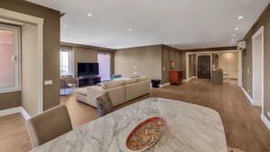 A spacious living area with a marble dining table, a beige sofa, and a television in the background.