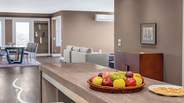 A modern kitchen with a marble countertop, a fruit basket, and a dining area with a table and chairs.
