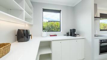 Et moderne køkken med hvide skabe, indbygget ovn, kaffemaskine og et vindue med udsigt til grøn natur.