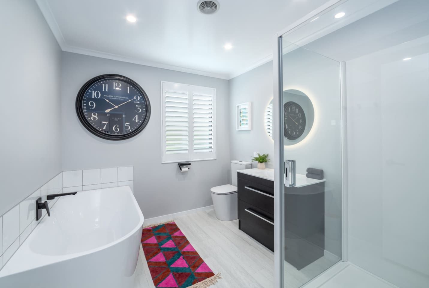A modern bathroom with a freestanding bathtub, a round wall clock, a toilet, and a glass shower enclosure.