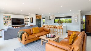 A modern living room with a sofa, coffee table, and a TV. There is a kitchen area with a dining set and a window with a view. - Sotally Tober Luxury Retreat | New Build, Spa, Walk to Square (Martinborough)