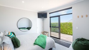 A modern bedroom with a large bed, a round mirror, a flat-screen TV, and a sliding glass door leading to a balcony with a view of greenery. - Sotally Tober Luxury Retreat | New Build, Spa, Walk to Square (Martinborough)