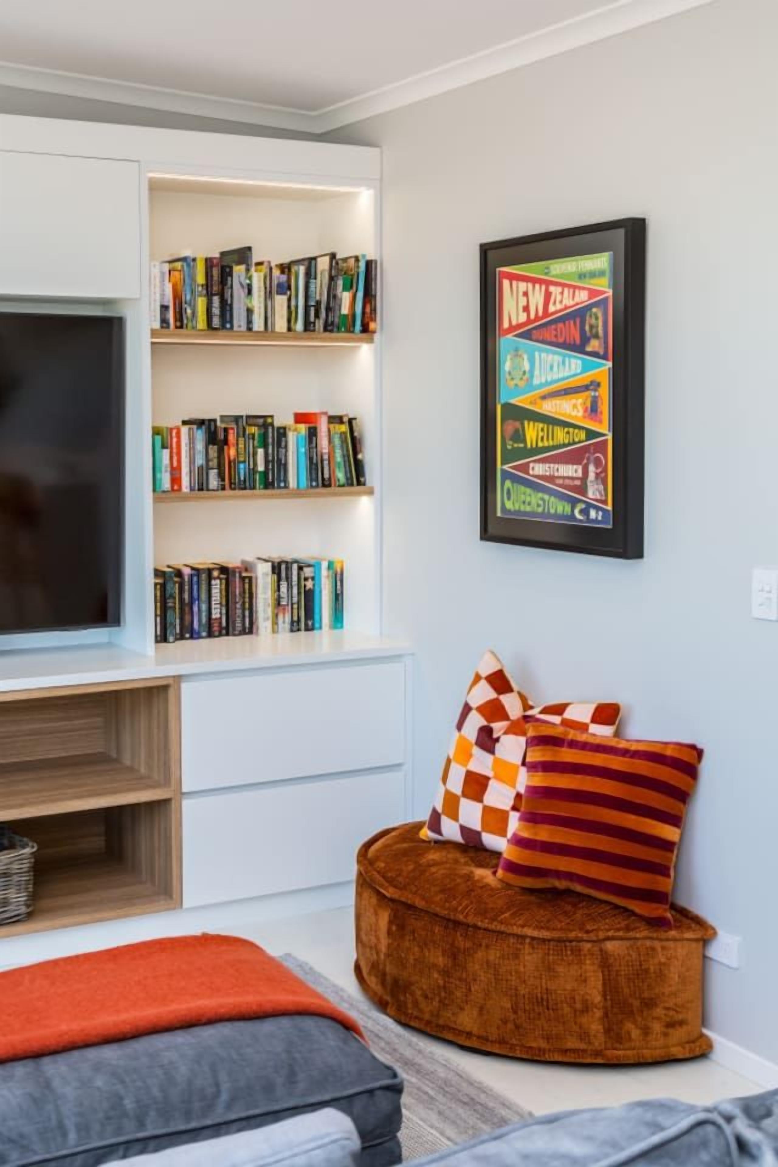 A modern living room with a bookshelf, a framed travel poster, and a round ottoman.