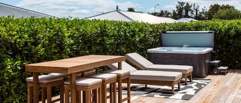 Una terraza exterior con una mesa de madera y sillones, un jacuzzi y un seto al fondo.