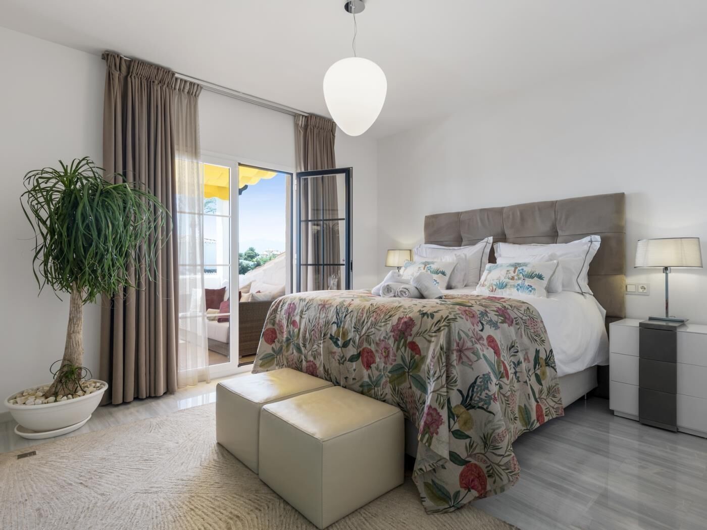 A modern bedroom with a large bed, a bedside table, a nightstand, a potted plant, and a view through a sliding door.