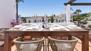 A dining table set outdoors with wicker chairs, a wine bottle, and a view of white buildings in the background. - Apartment in Aldea Blanca for 6 persons |Nueva Andalucía (Nueva Andalucía)