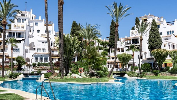 A swimming pool surrounded by white buildings with balconies, palm trees, and a clear blue sky. - Apartment in Aldea Blanca for 6 persons |Nueva Andalucía (Nueva Andalucía)