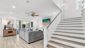 Ein geräumiges Wohnzimmer mit grauem Sofa, Holzboden, Deckenventilator und Treppe mit weißen Geländern.