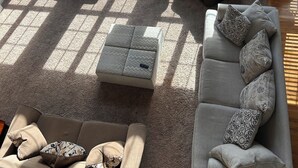 Ein geräumiges Wohnzimmer mit großen Fenstern, einem bequemen Sofa, einem Sessel und einem kleinen Hocker.