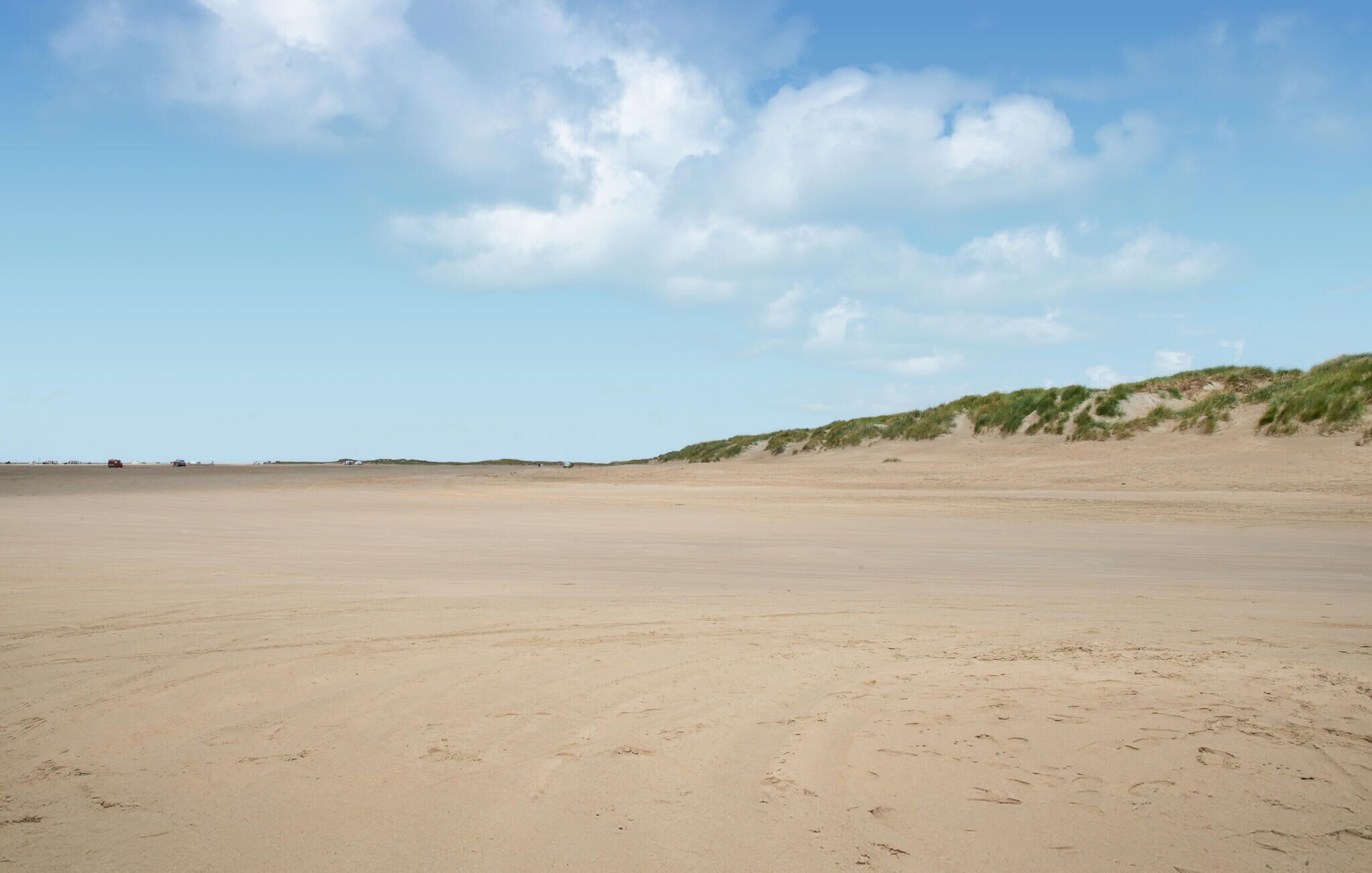 Een breed, open zandstrand met duinen op de achtergrond.