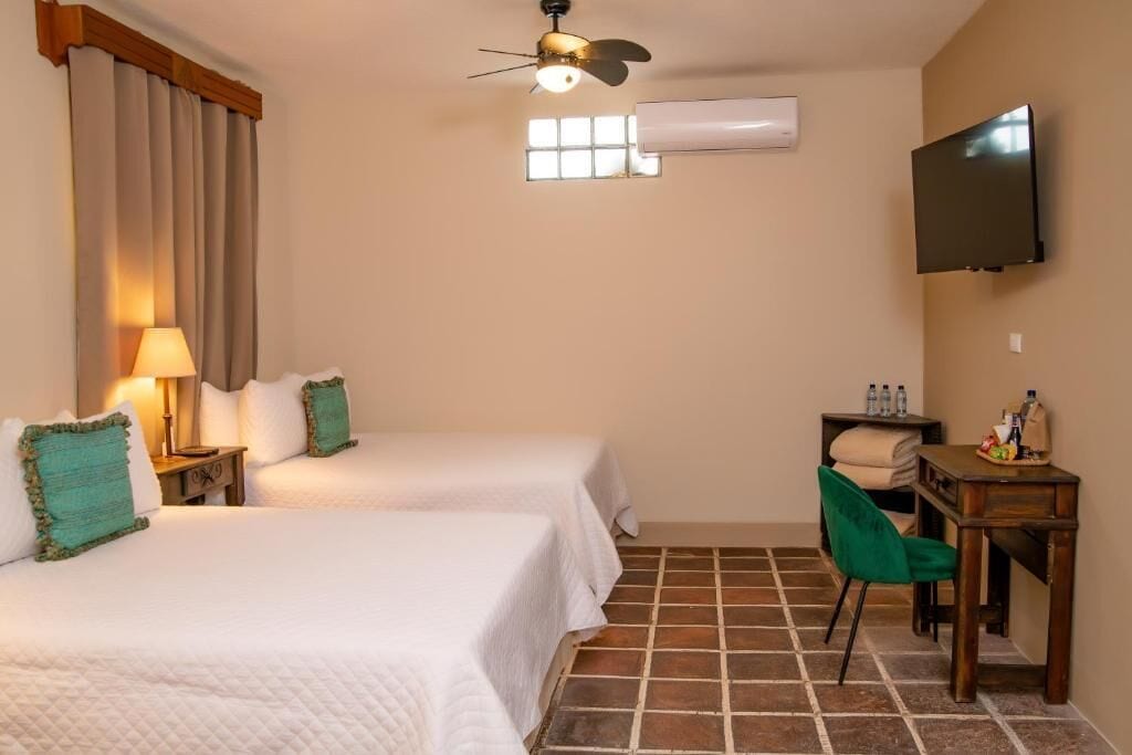 A hotel room with two beds, a desk, a chair, a TV, an air conditioner, and a window with curtains.