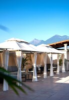 Un espacio al aire libre cubierto con mesas y sillas, un sofá, y vistas a montañas al fondo.