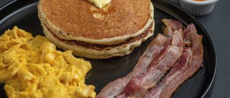 A breakfast plate with pancakes, scrambled eggs, bacon, and a fruit bowl.