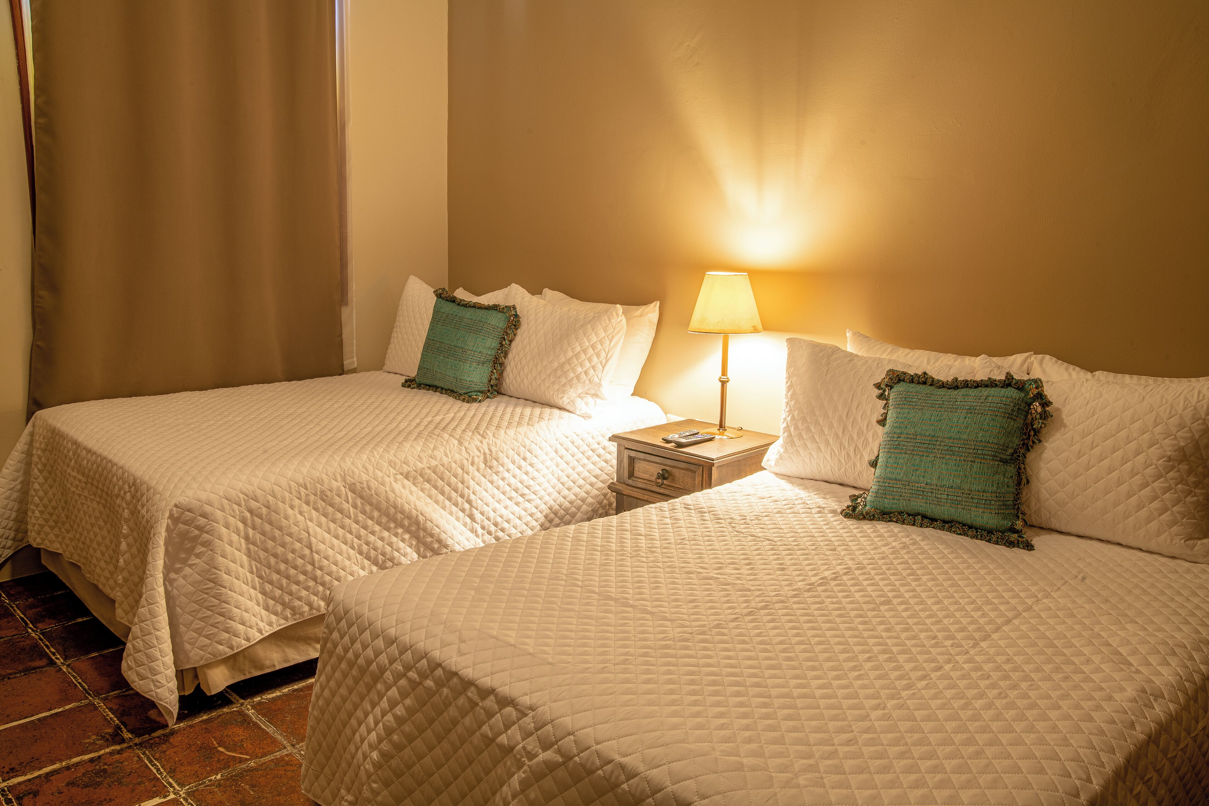 Two beds with white quilts and green pillows, a nightstand with a lamp, and a tiled floor.