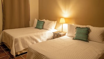 Two beds with white quilts and green pillows, a nightstand with a lamp, and a tiled floor.