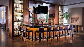 A bar area with a long counter, multiple bar stools, shelves with liquor bottles, a flat-screen TV, and a wooden barrel.