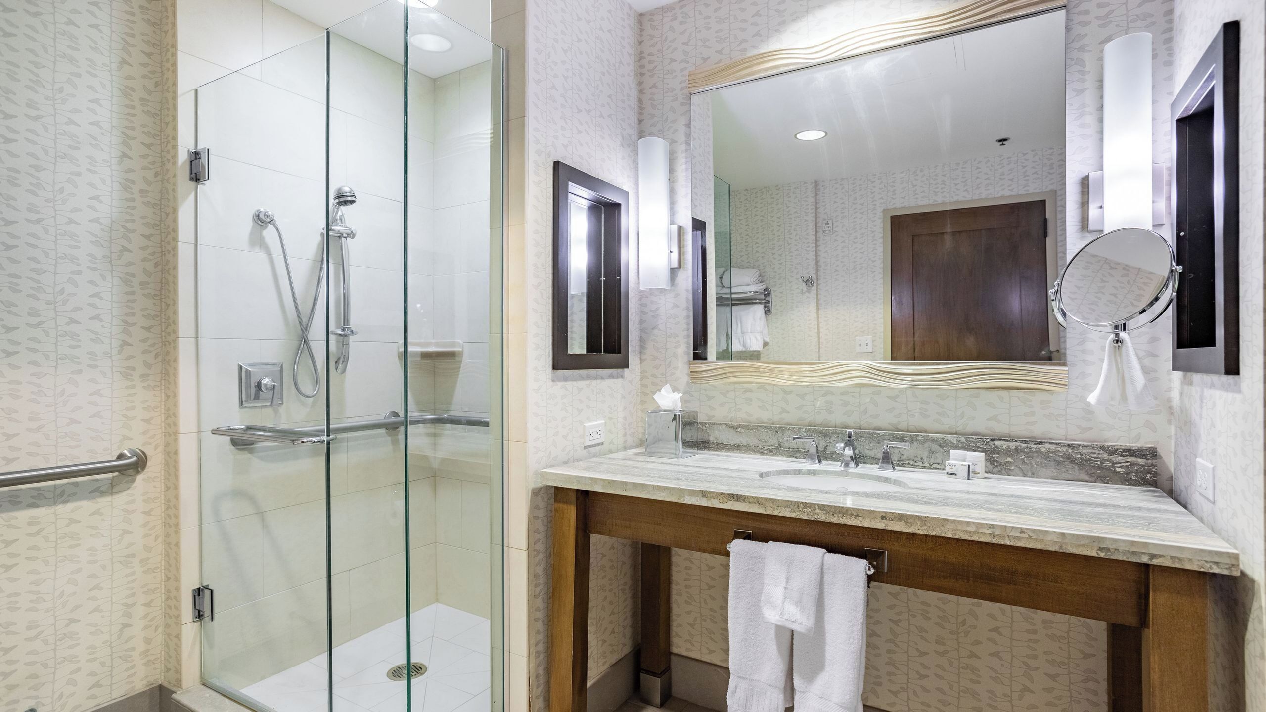 A hotel bathroom with a glass-enclosed shower, a large mirror, and a marble sink.