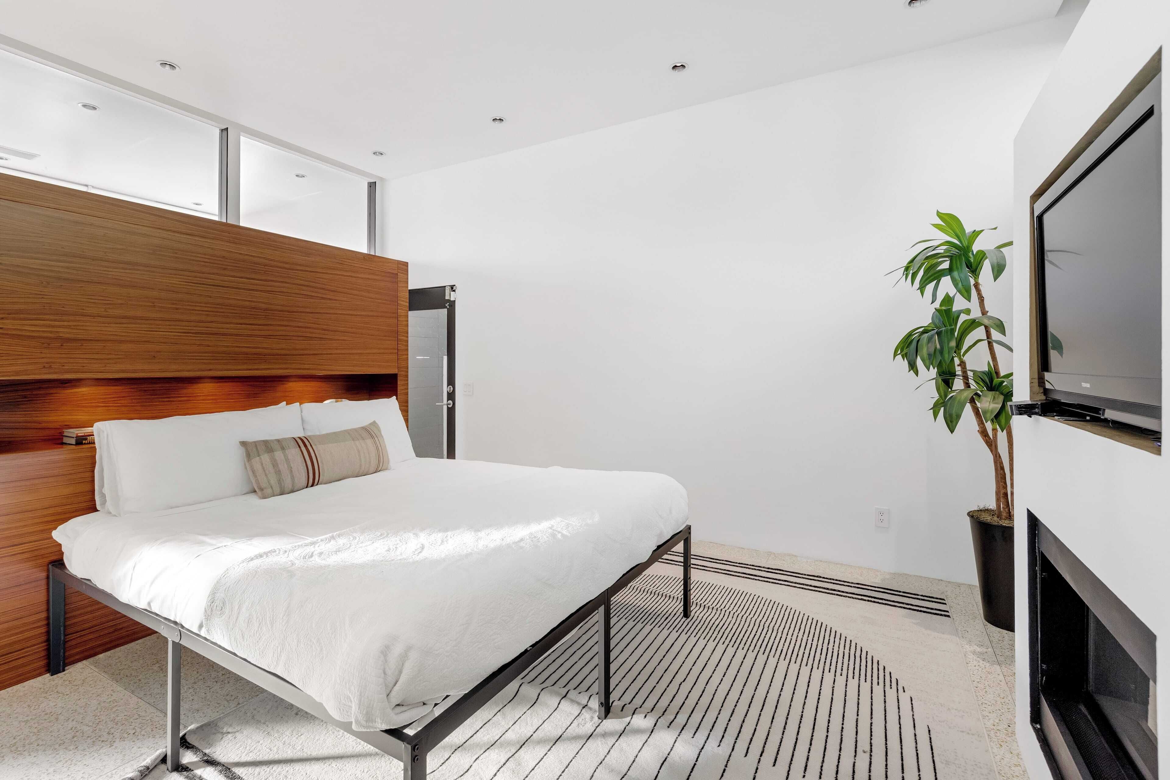 Un dormitorio moderno con cama, cabecero de madera, planta en maceta y televisor montado en la pared.